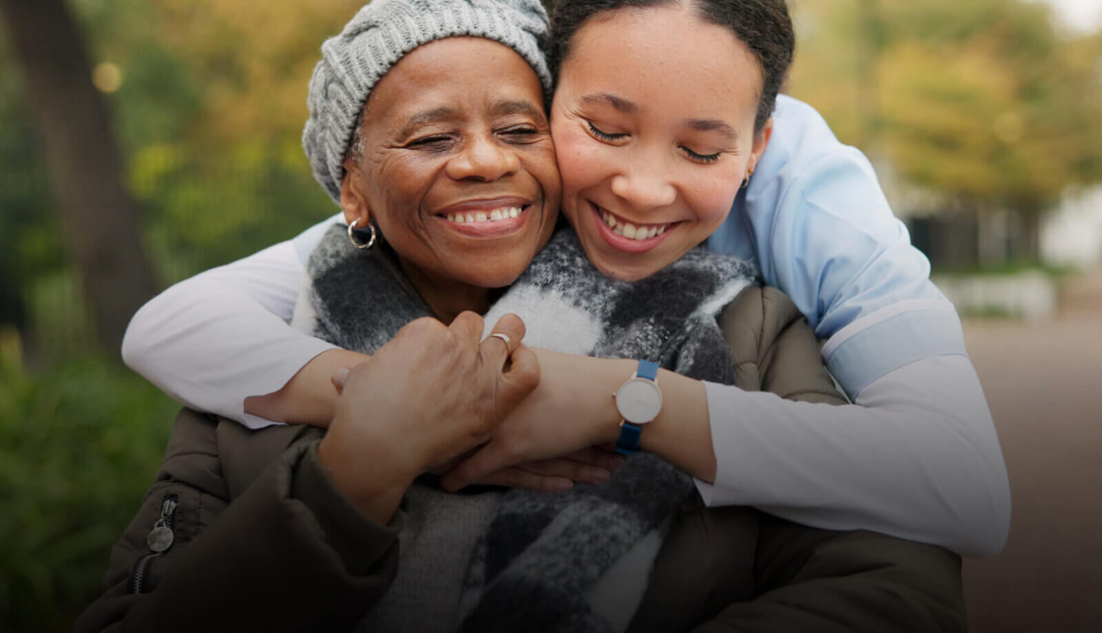 Young person hugging an older person who is in a wheelchair. They are both outside and are smiling.