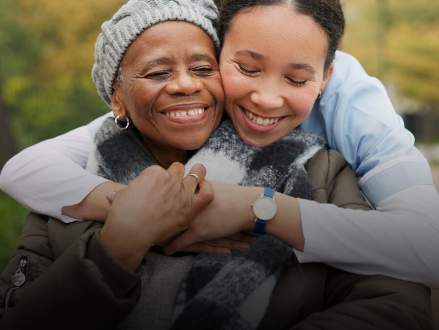 Young person hugging an older person who is in a wheelchair. They are both outside and are smiling.